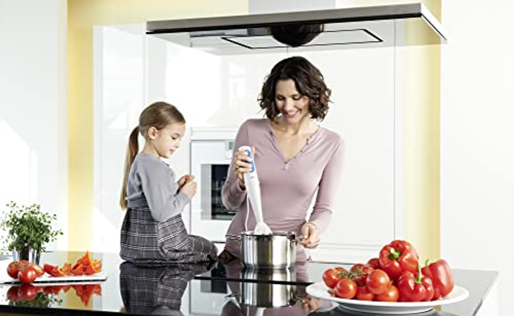 Eine Frau und ein Mädchen in einer Küche benutzen einen Stabmixer mit einem Topf mit Essen und Gemüse.