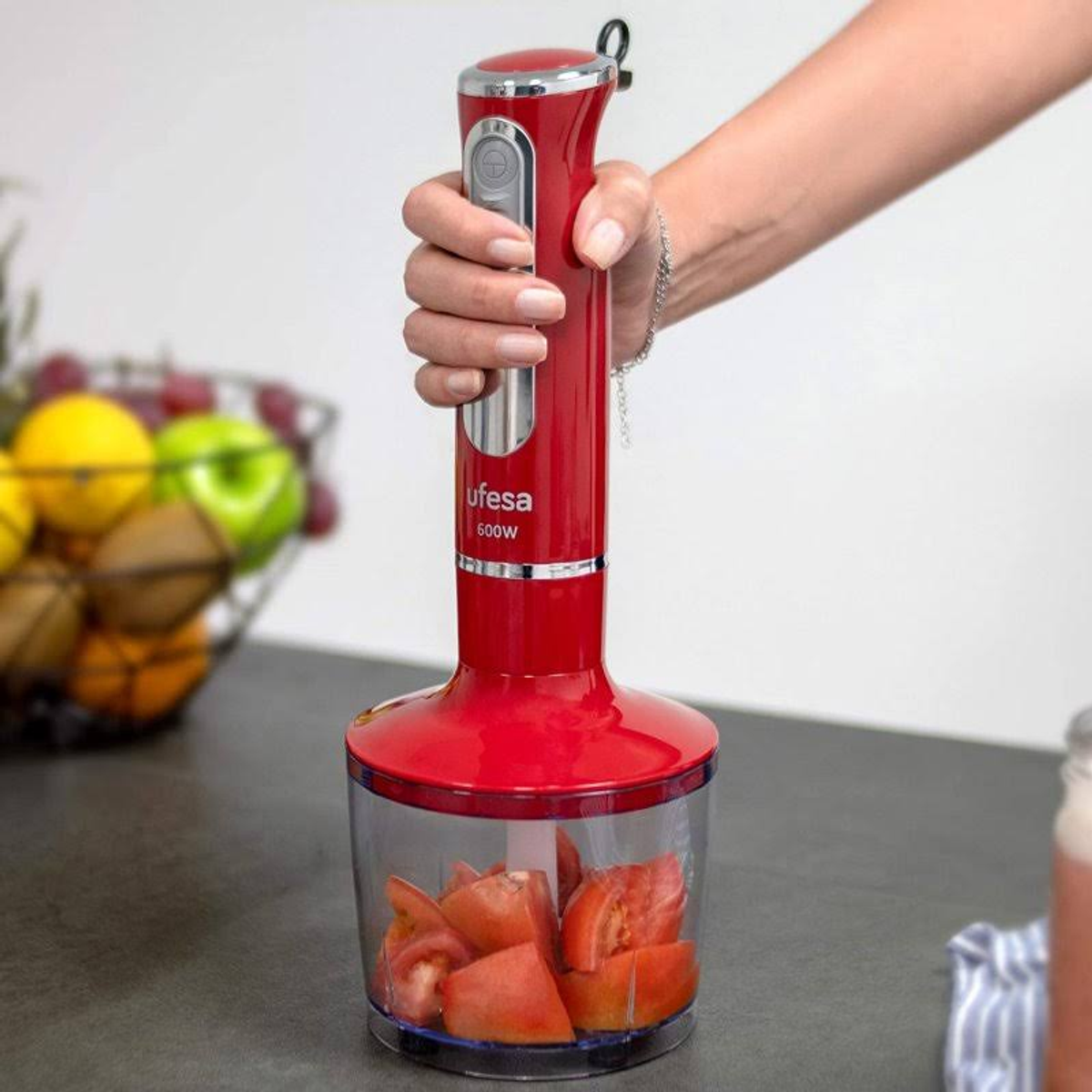 Un frullatore a immersione rosso con pomodori in un contenitore, tenuto da una donna. Cesto di frutta.