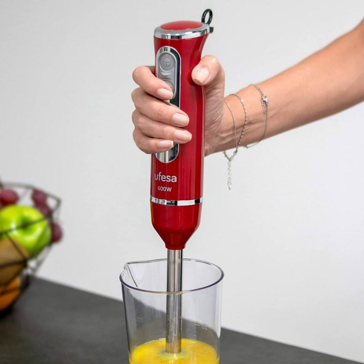 Un frullatore a immersione rosso in uso, che frulla liquido giallo in un becher trasparente. Si vede una mano.