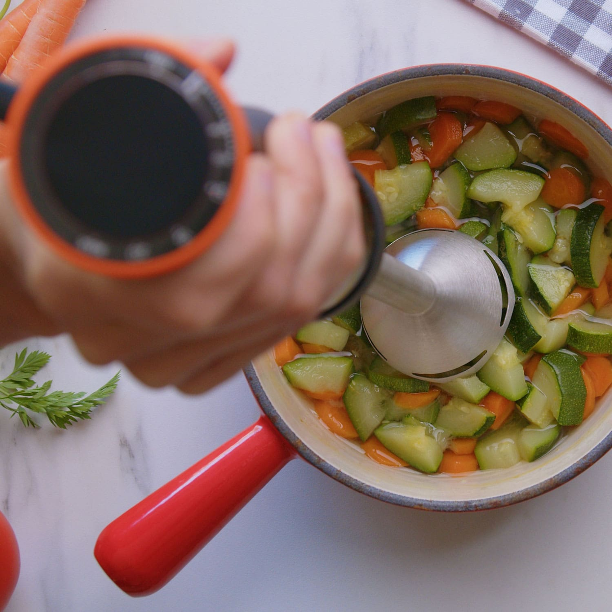 Ręka używająca blendera ręcznego w garnku z posiekanymi warzywami i bulionem.
