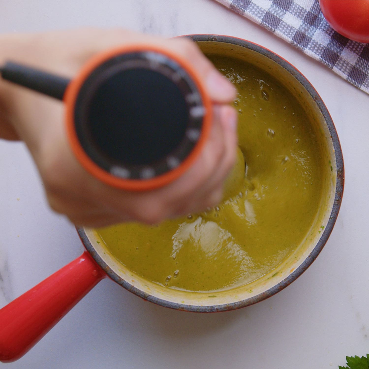 Ręka trzyma blender w czerwonym garnku z zieloną zupą. Ręcznik i pomidor w tle.