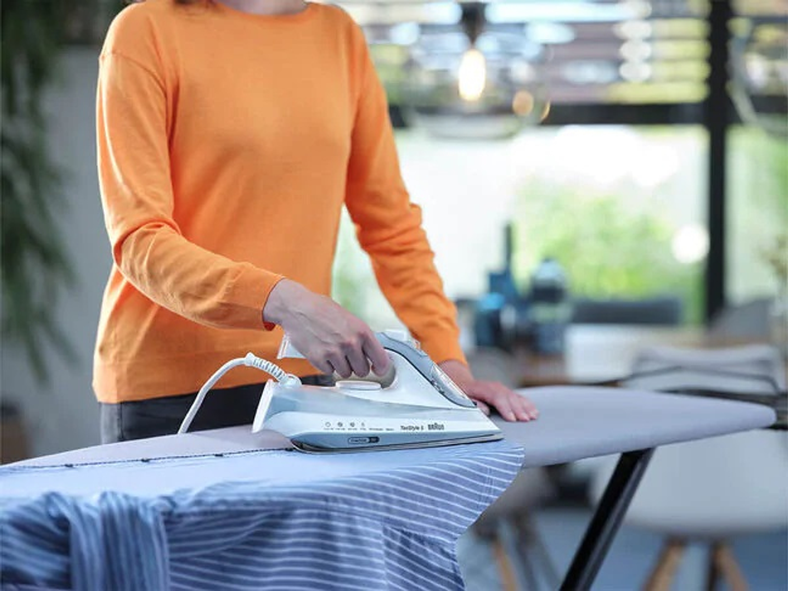 Personne repassant une chemise rayée bleue et blanche sur une planche à repasser. La personne porte une chemise orange.