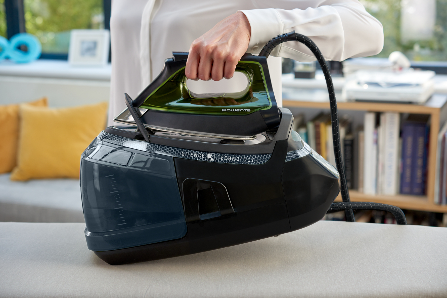 Eine Person hält ein schwarzes Bügeleisen auf einem weißen Bügelbrett. Das Bügeleisen befindet sich auf einer schwarzen Basis mit einem Wassertank.
