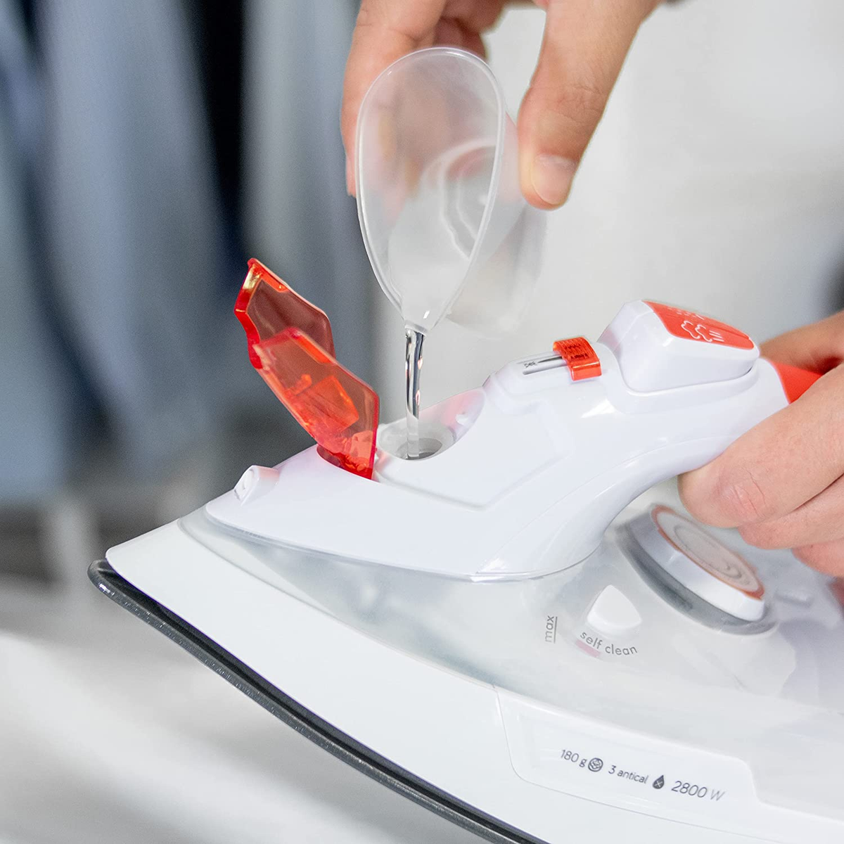 Eine Person gießt Wasser in ein weiß-rotes Bügeleisen. Details auf dem Bügeleisen sind max. Füllstand und Selbstreinigung.