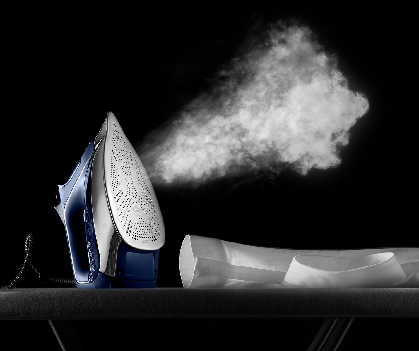 Plancha con vapor. Plancha azul y plateada sobre una superficie negra. Sale vapor. Fondo oscuro.
