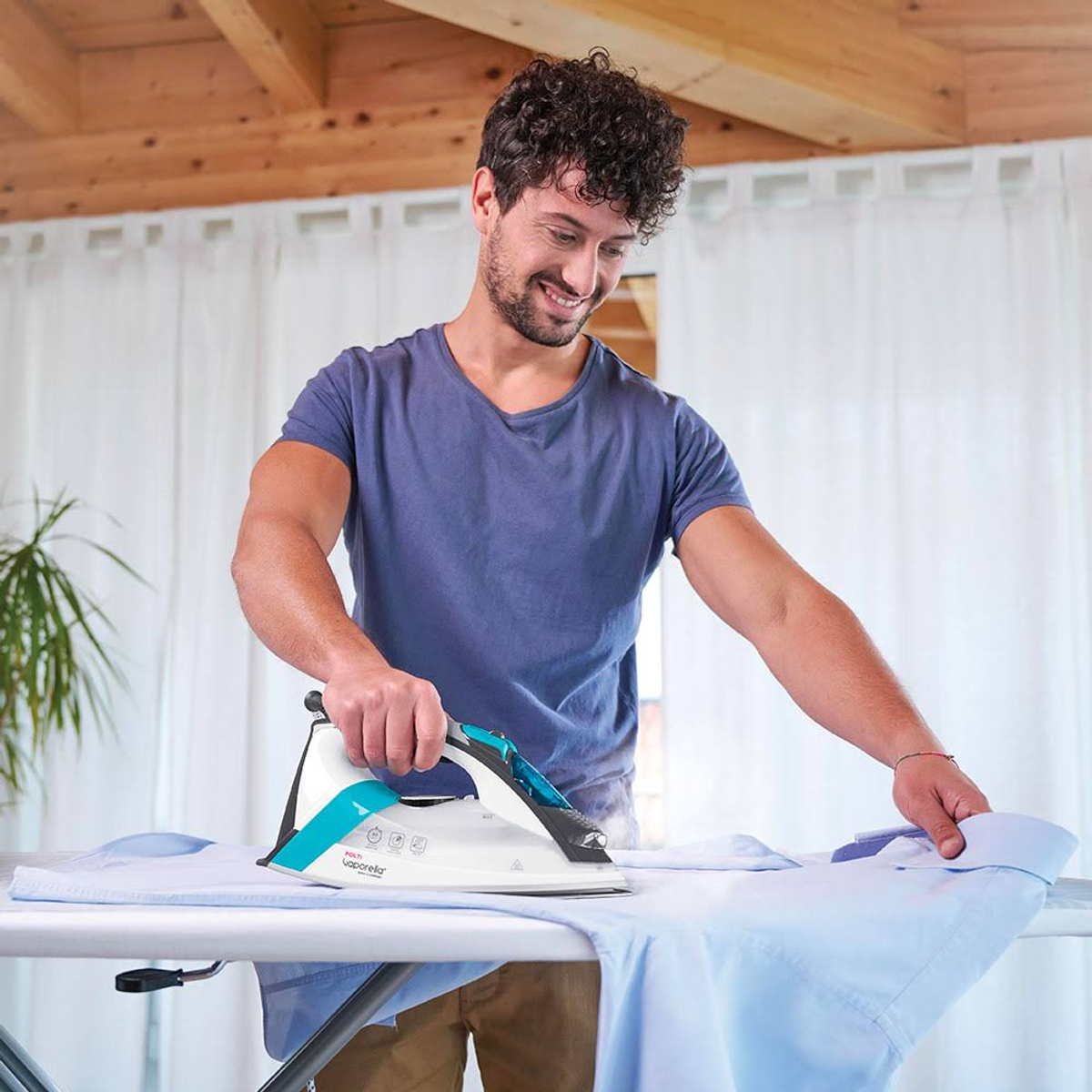 Mężczyzna prasujący niebieską koszulę na desce do prasowania białym i turkusowym żelazkiem.