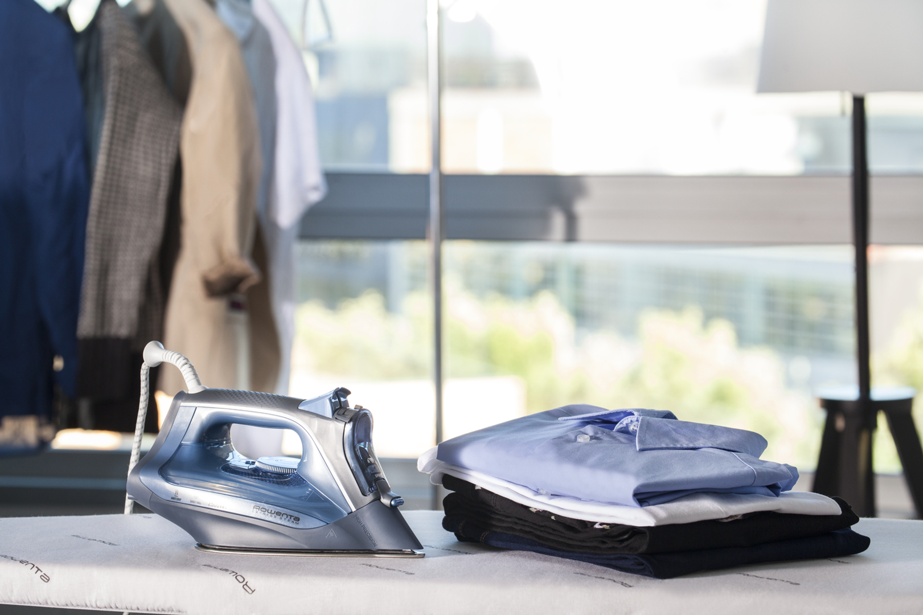 Deska do prasowania z żelazkiem i stosem ubrań. Tło ma okno i trochę ubrań.