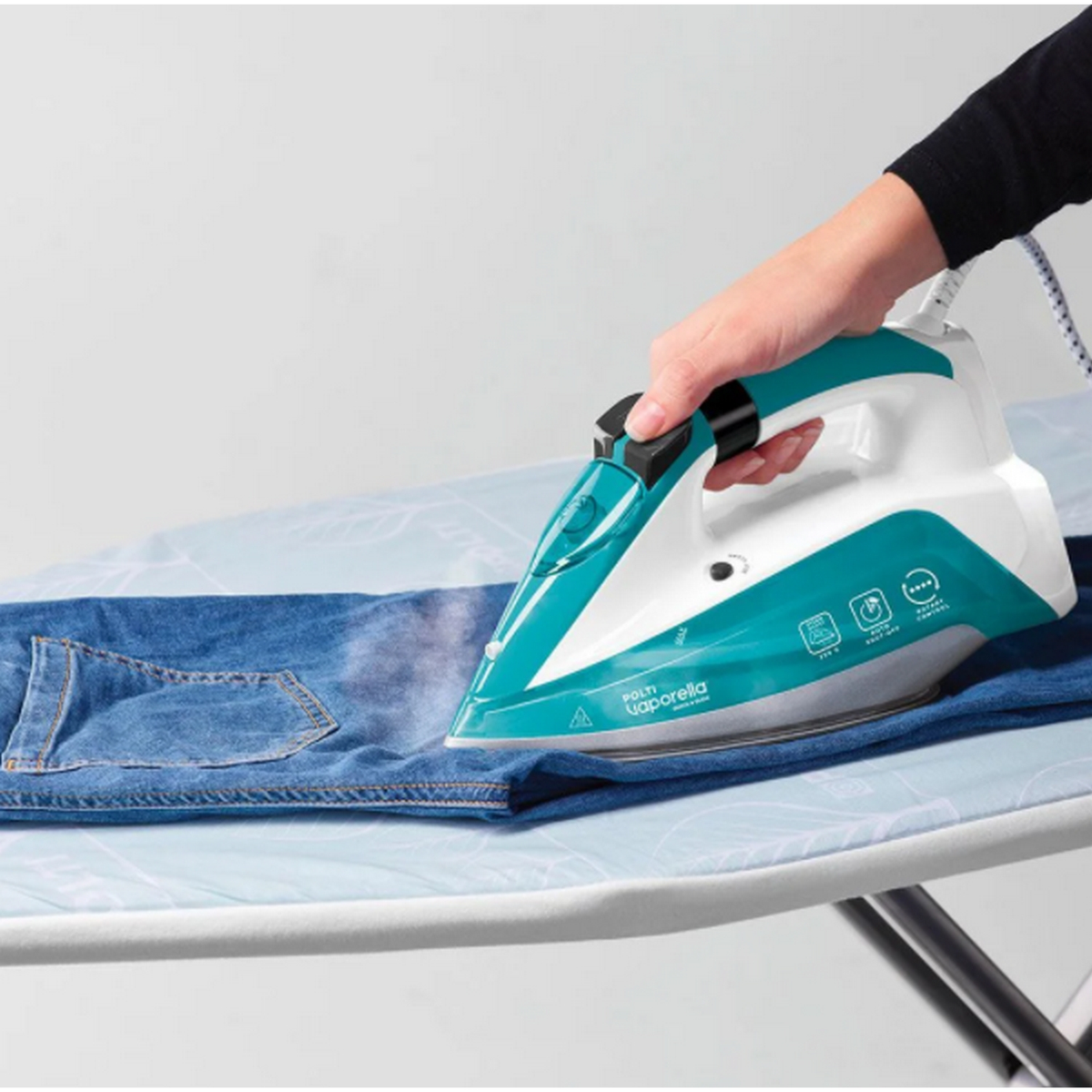 Osoba prasuje niebieskie dżinsy na biało-niebieskiej desce do prasowania. Para jest widoczna.