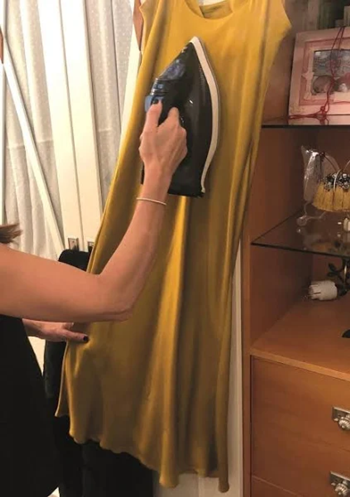 Una persona planchando un vestido amarillo. La plancha es negra y el vestido cuelga de una pared.