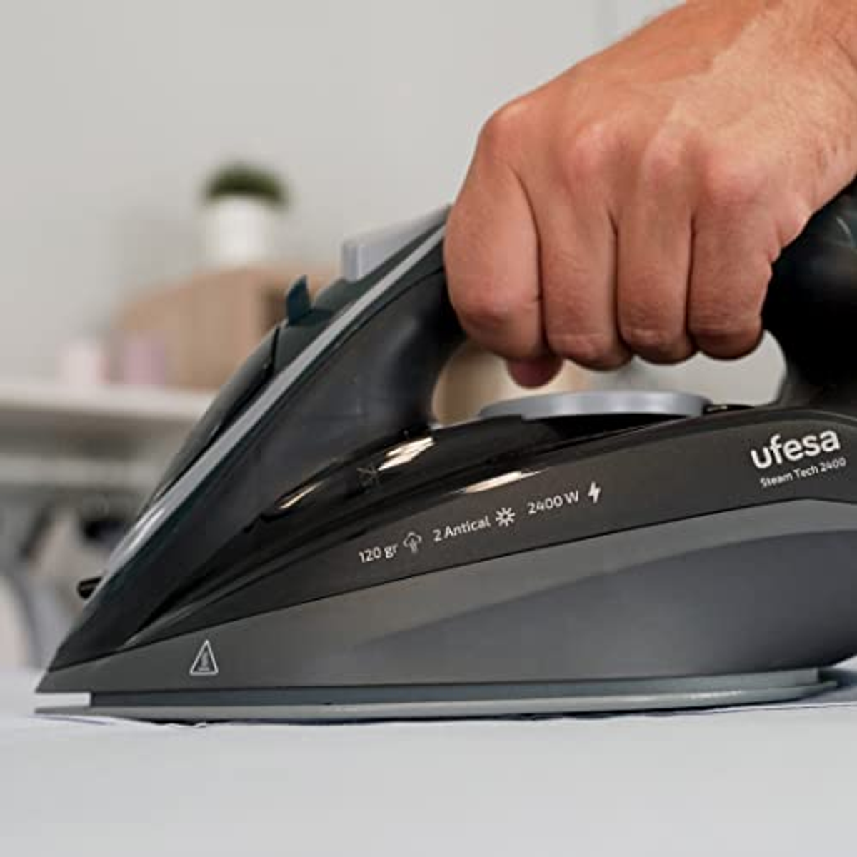 Die Hand einer Person bügelt ein Kleidungsstück mit einem dunkelgrauen Bügeleisen. Das Bügeleisen hat eine Beschriftung.