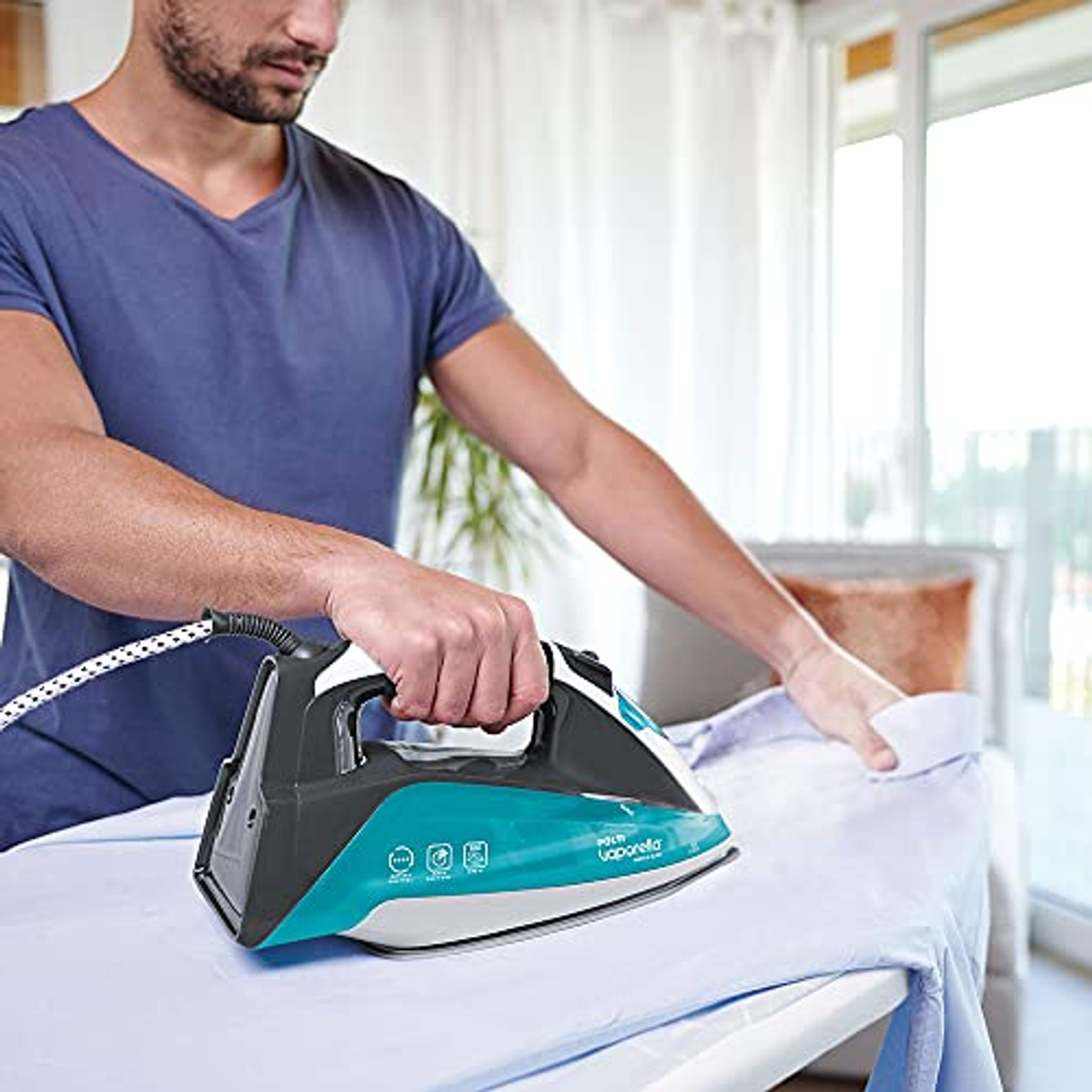 Mężczyzna prasujący koszulę. Żelazko jest czarno-turkusowe, marki 'Polti'. Mężczyzna ma niebieską koszulę.