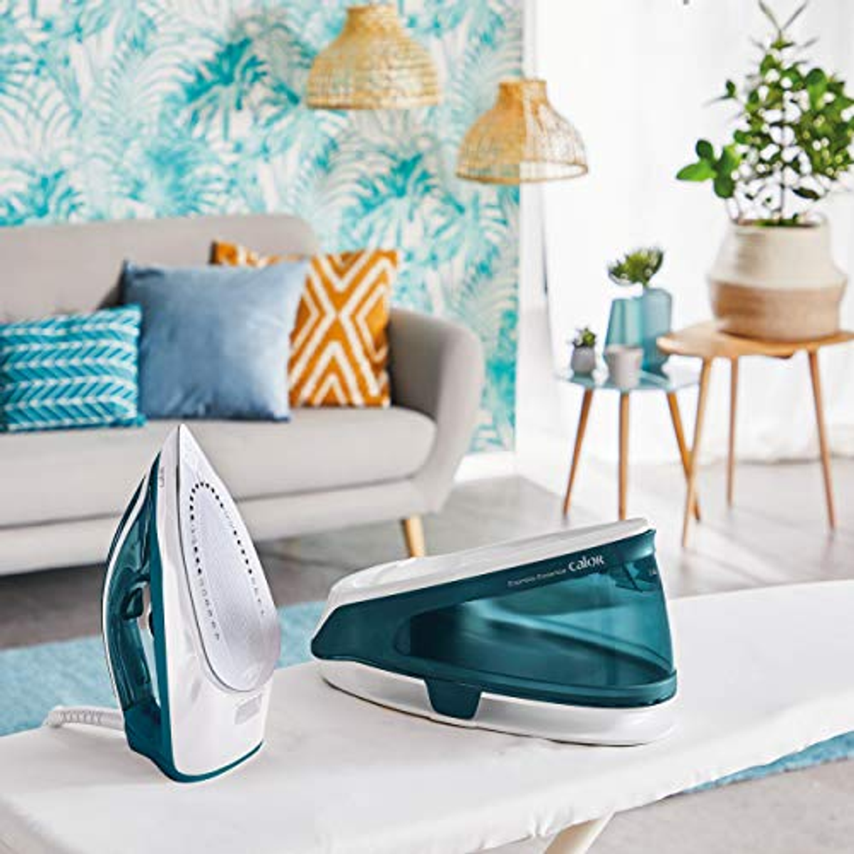 Tabla de planchar con dos planchas. La habitación tiene papel pintado floral azul y blanco, sofá y plantas.