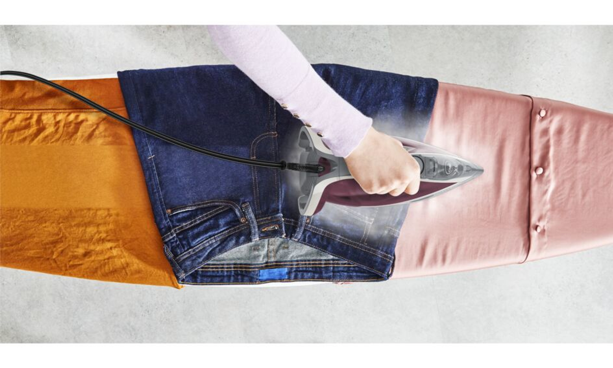 Eine Person bügelt blaue Jeans auf einem Bügelbrett. Das Bügeleisen gibt Dampf ab.