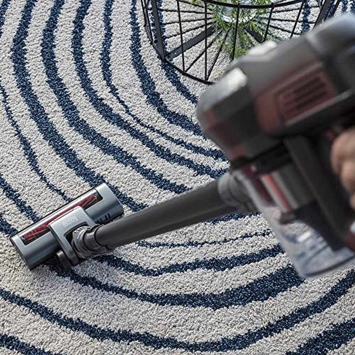 Eine Hand benutzt einen Staubsauger auf einem Teppich. Teppich ist weiß mit blauen Streifen. Der Staubsauger ist grau.