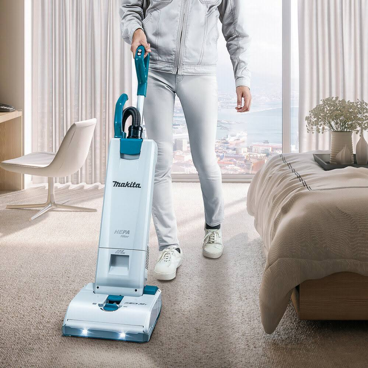 Eine Person saugt einen Raum mit einem weiß-blauen Staubsauger in der Nähe eines Bettes.