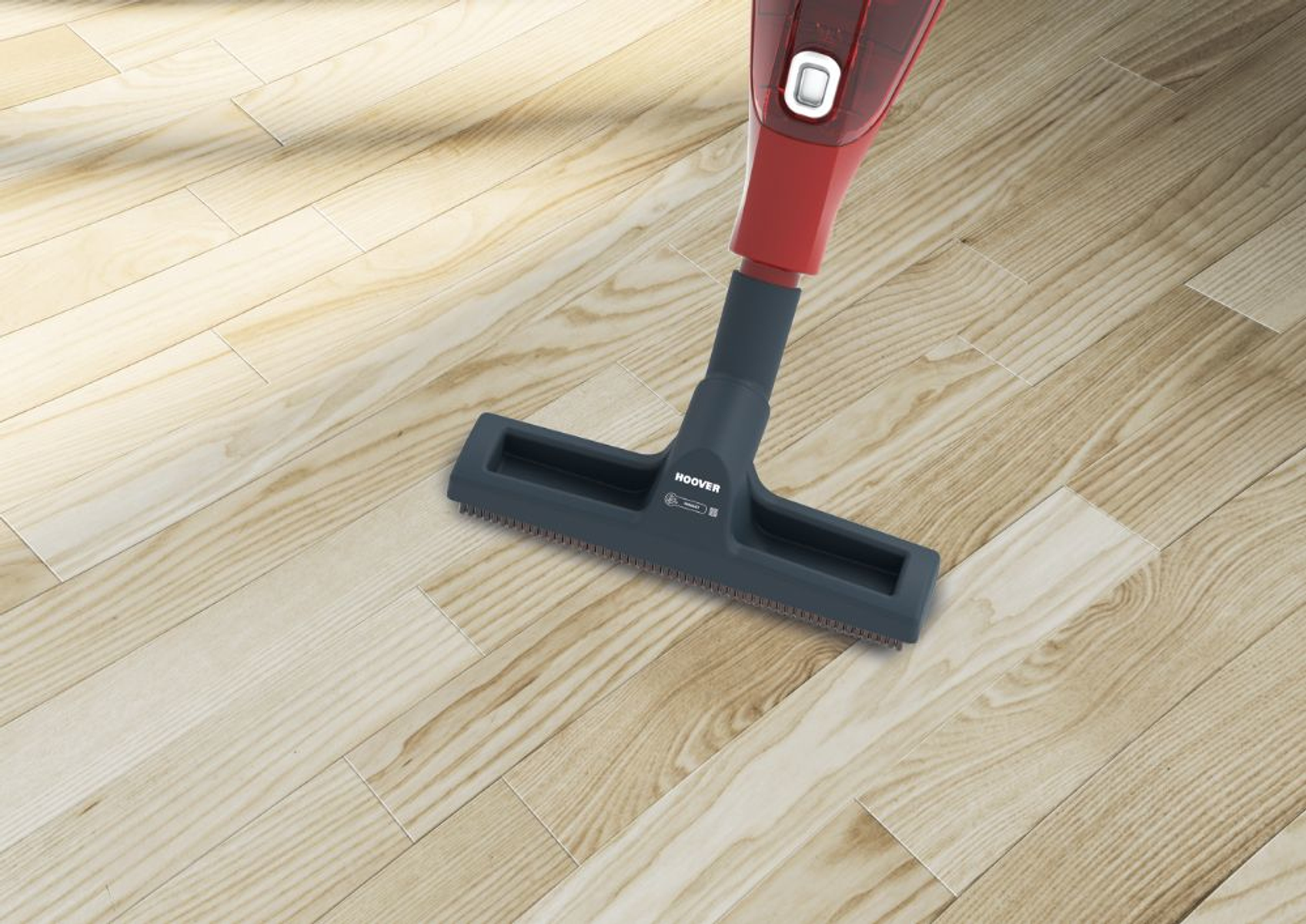 Una spazzola per aspirapolvere rossa e nera pulisce un pavimento in legno con venature visibili.