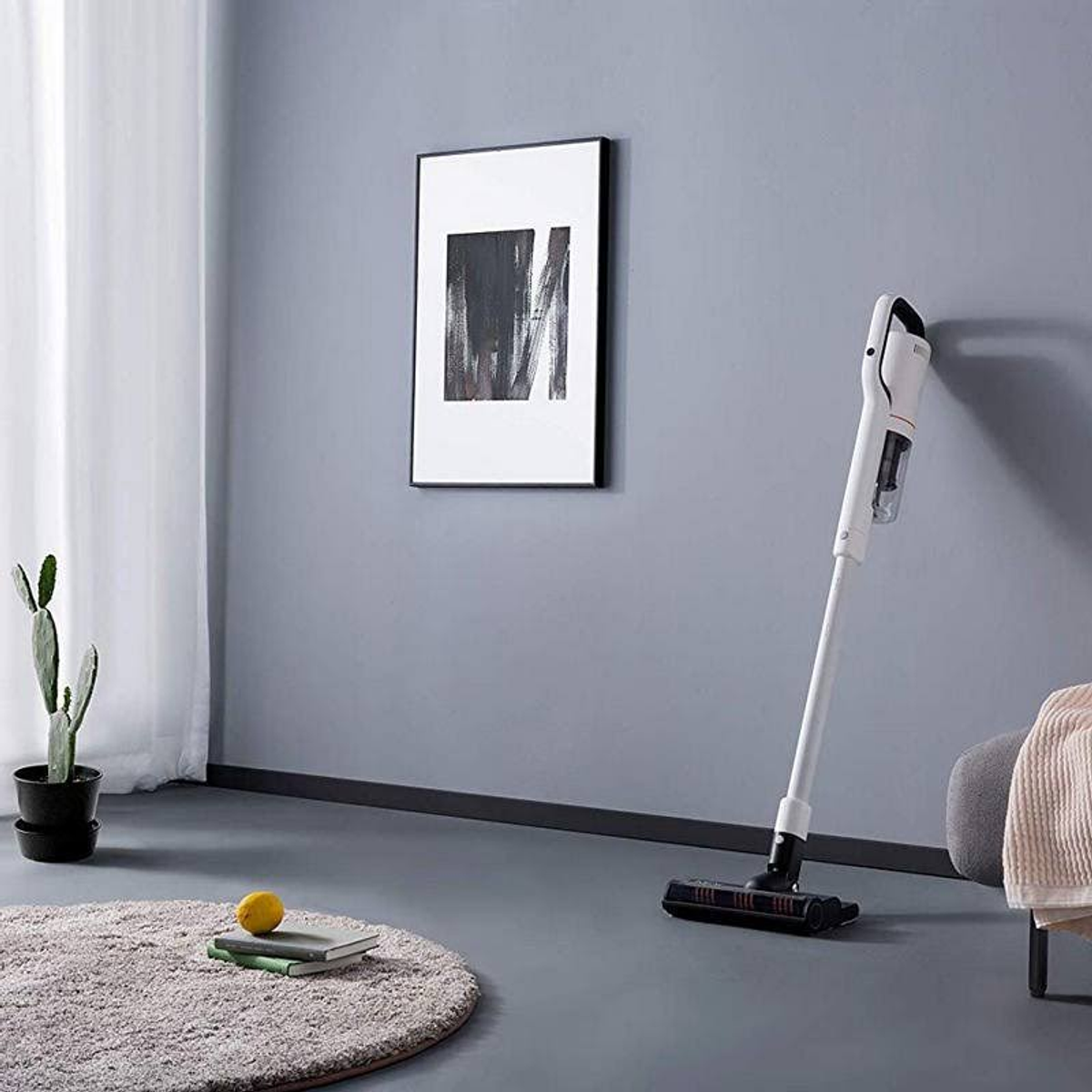 Aspirapolvere bianco e grigio appoggiato a parete grigia, arte e piante vicine.