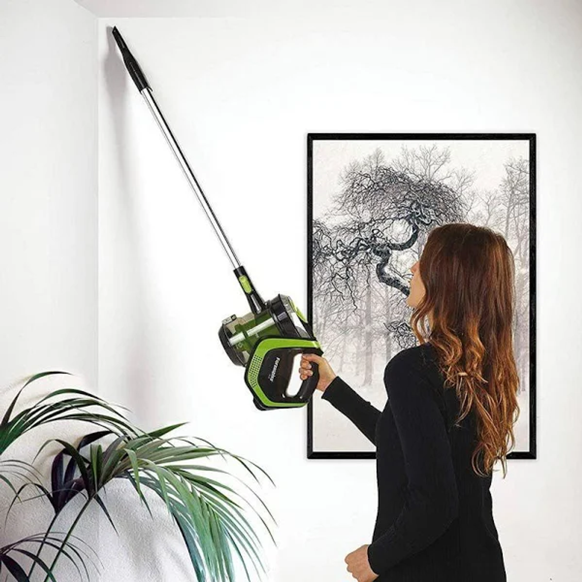 Una mujer aspira una pared con una aspiradora verde y negra, cerca de una planta y un cuadro.