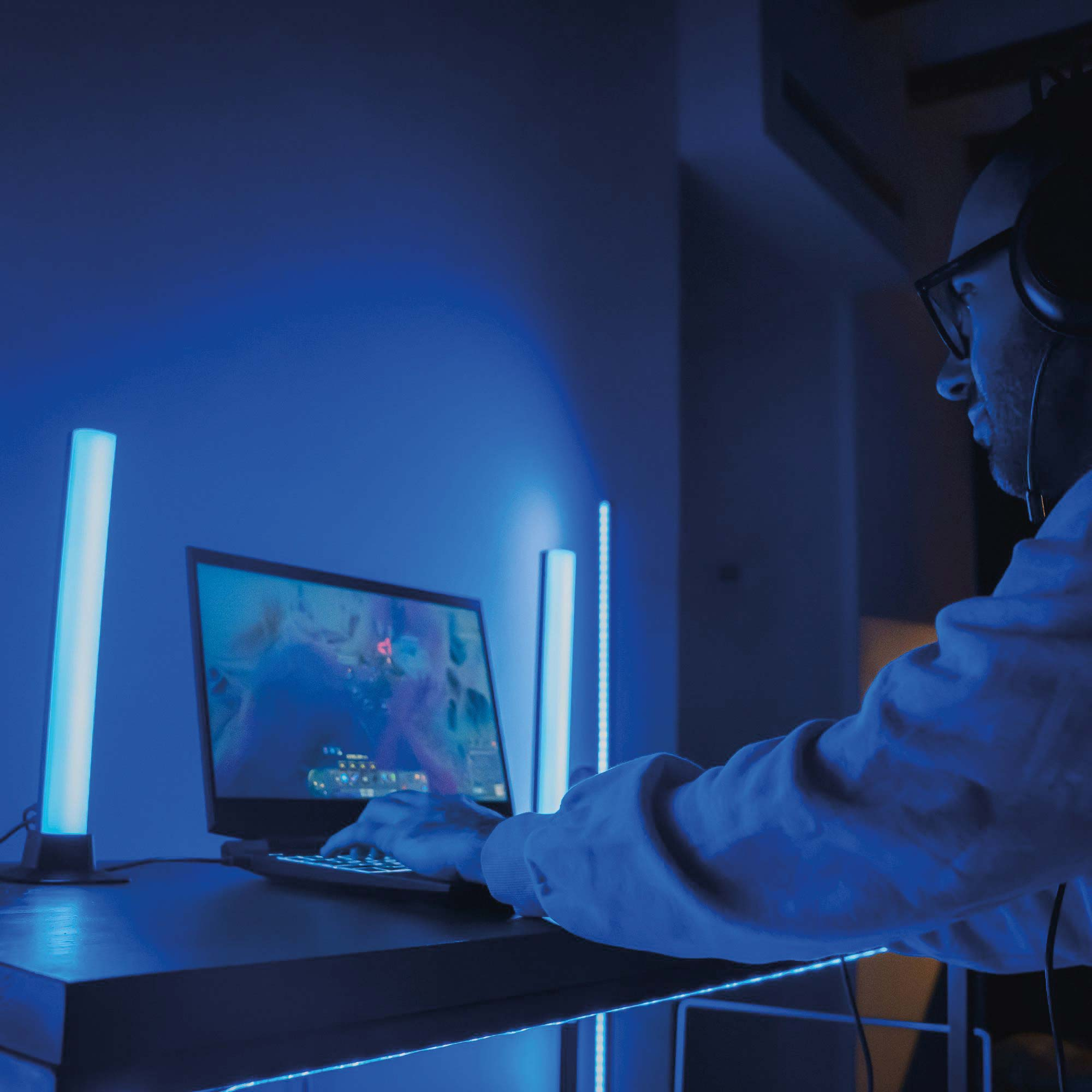 Mężczyzna gra na laptopie, niebieskie światła na ścianie, słuchawki. Ciemny pokój.