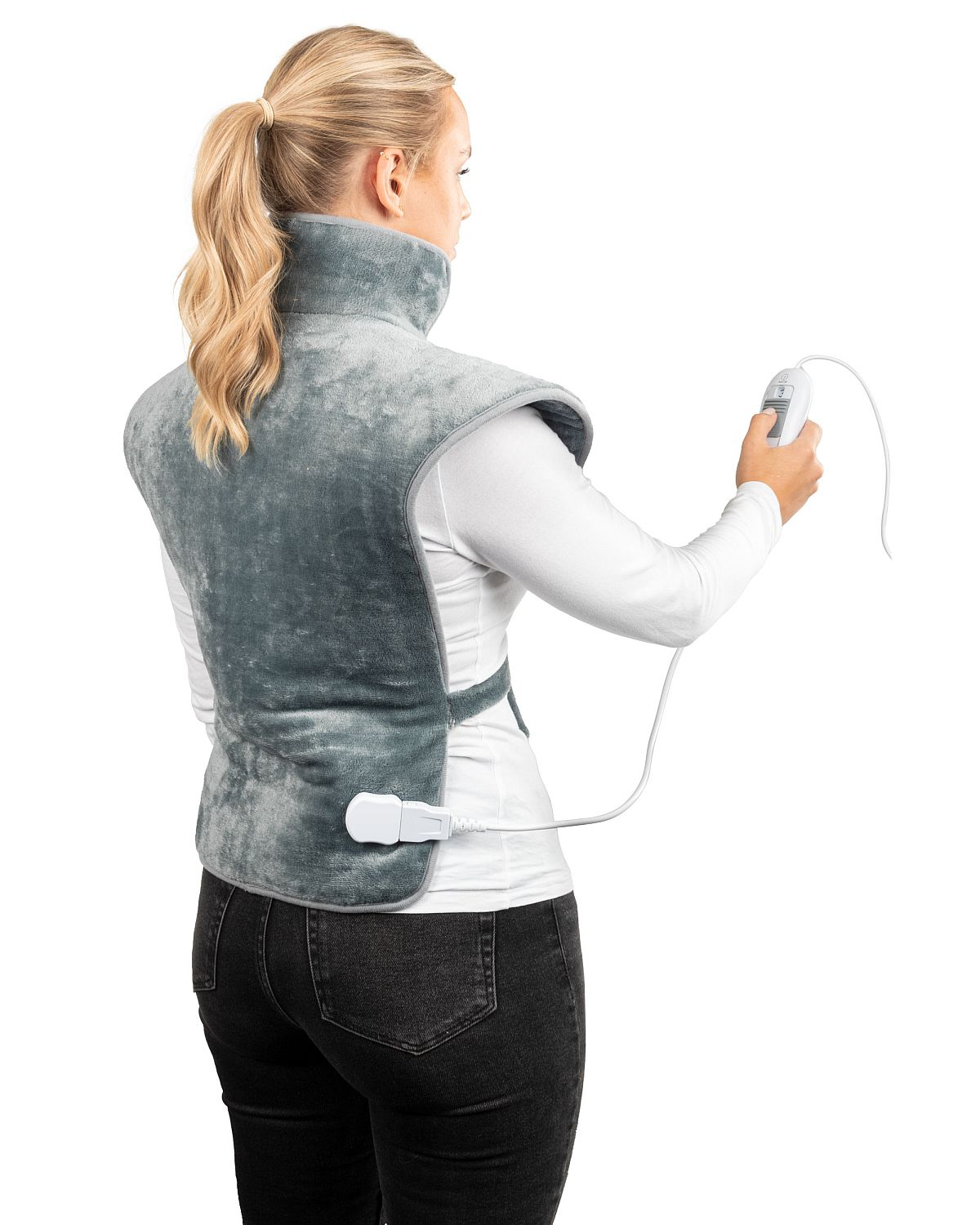 Una mujer con una almohadilla térmica gris alrededor del cuello y los hombros, sosteniendo el dispositivo de control. Viste camisa blanca y vaqueros negros.