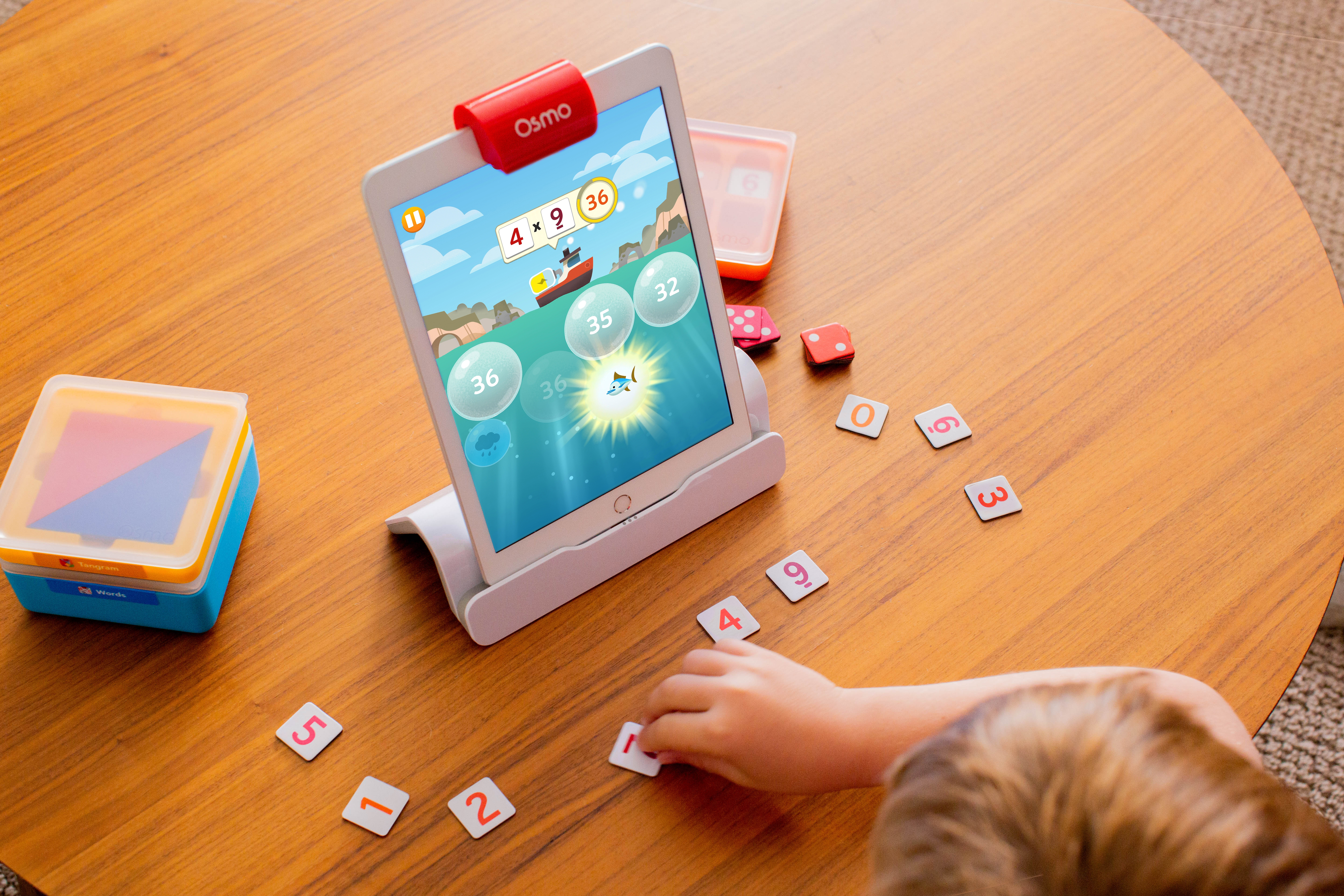 Bambino che gioca con tablet e numeri. Tablet mostra gioco di matematica. Tavolo di legno. Pezzi di gioco sparsi.