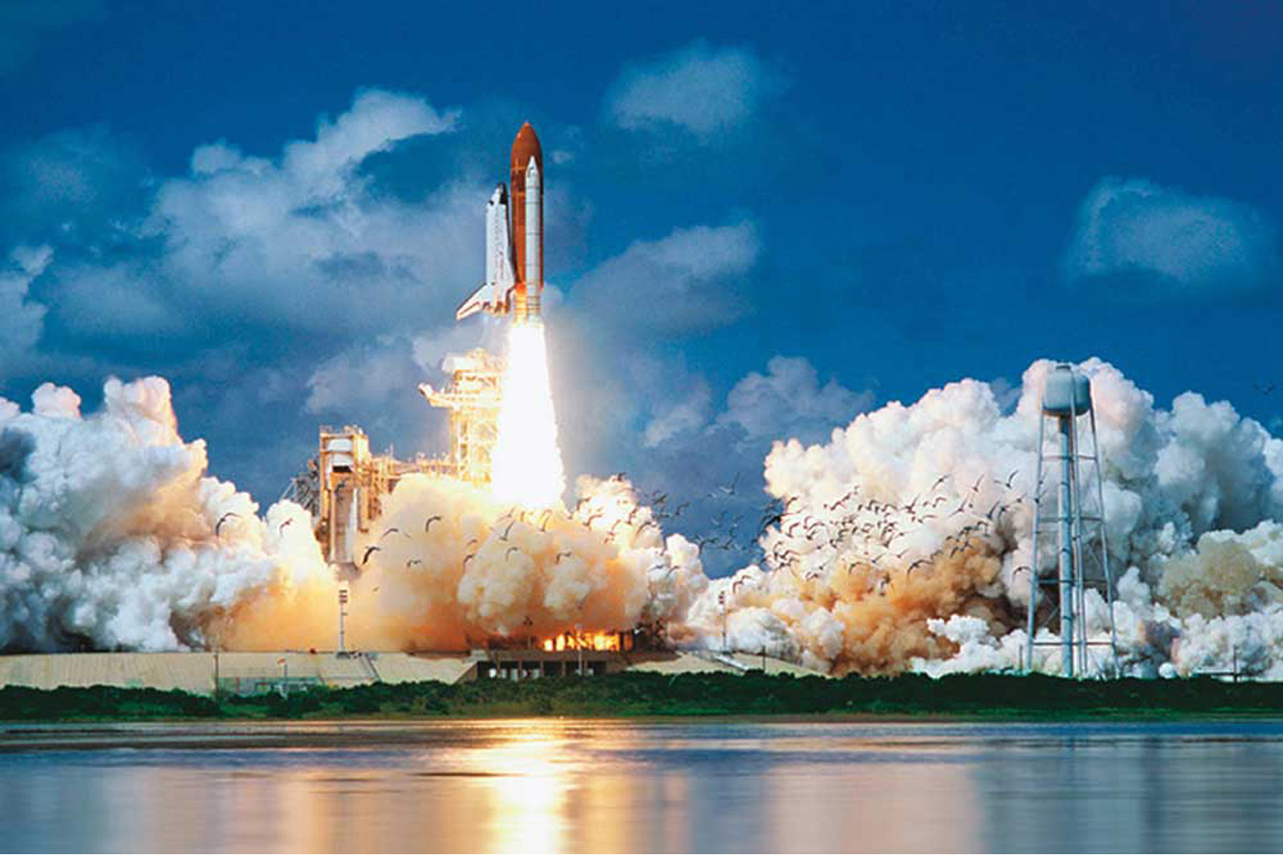 Ein Space Shuttle startet. Es ist von Rauch, Feuer und einem Wasserturm im Hintergrund umgeben.