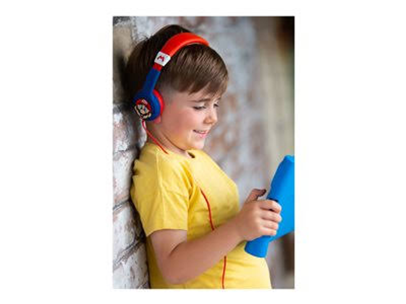 Chłopiec w czerwono-niebieskich słuchawkach, patrzący na niebieski tablet, oparty o ceglaną ścianę.