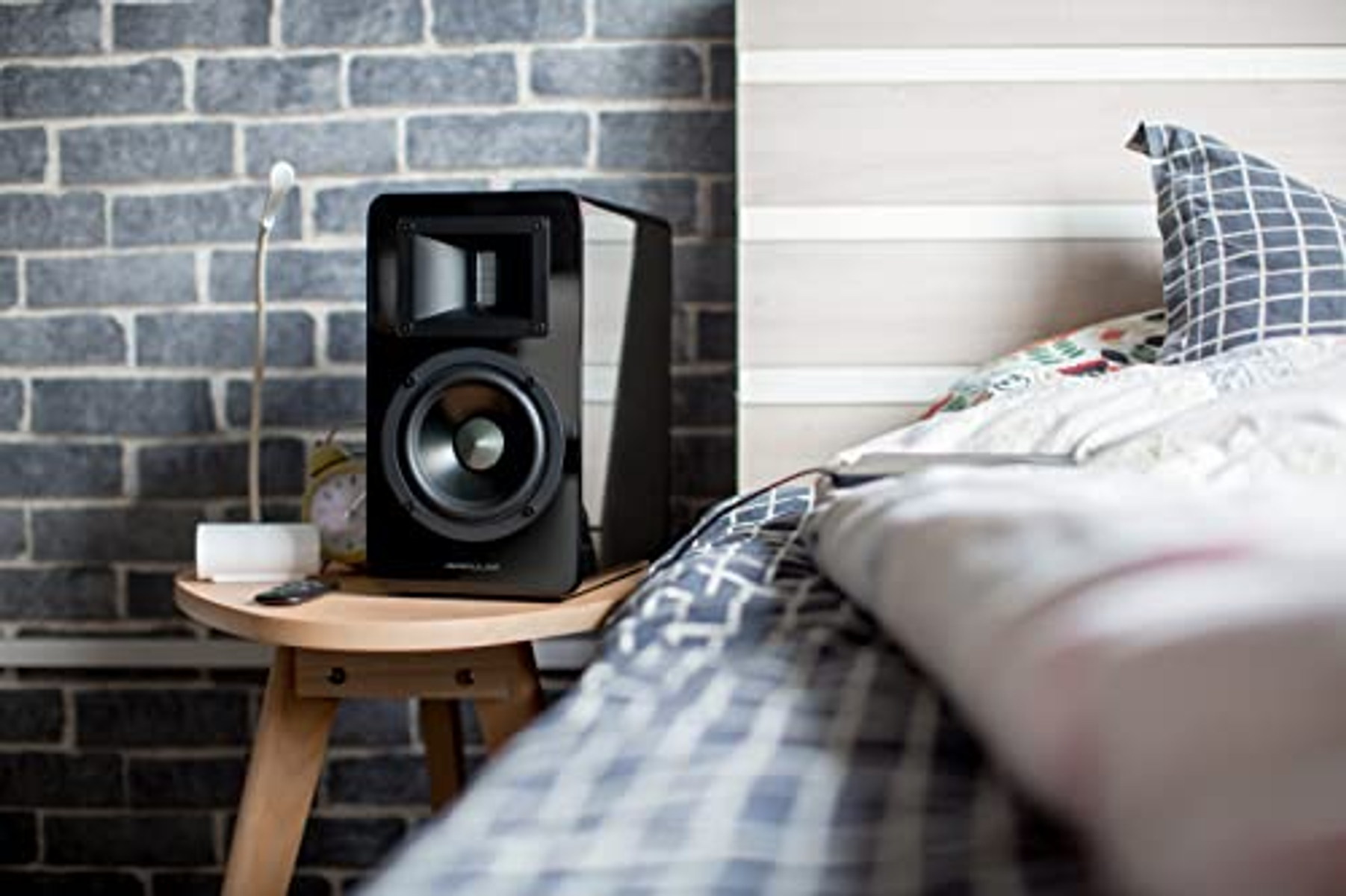 Czarny głośnik na drewnianym stoliku nocnym obok łóżka. W pobliżu lampa i pilot.