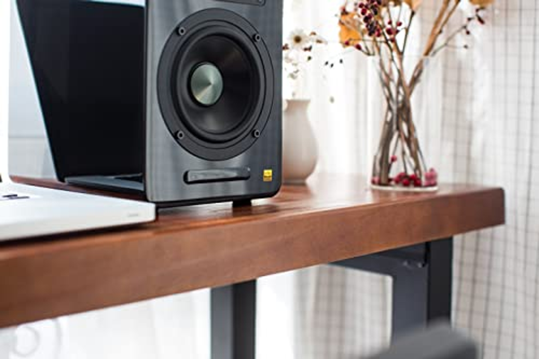 Głośnik na drewnianym biurku. Za nim laptop i wazon z kwiatami.