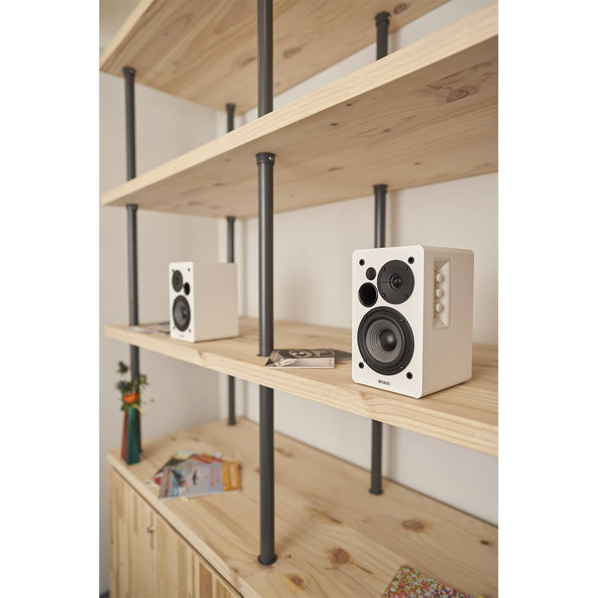 Dwa białe głośniki na drewnianej półce. Jeden bliżej, drugi dalej. Na półce kilka przedmiotów.