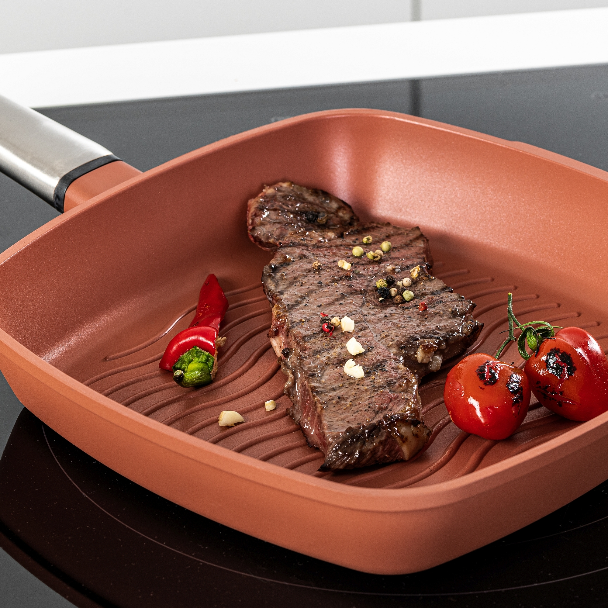 Gegrilltes Steak und Tomaten in einer kupferfarbenen Grillpfanne auf einem schwarzen Kochfeld.