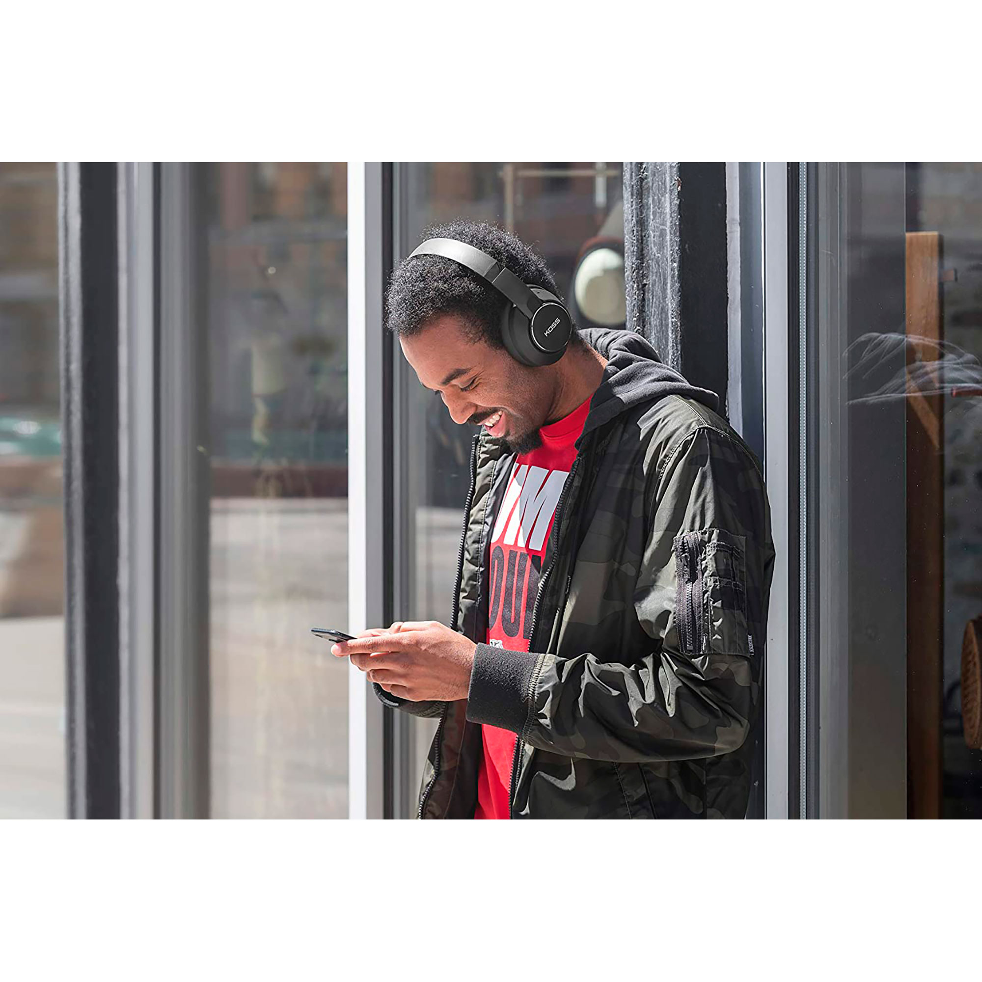 Un homme avec un casque regardant un téléphone. Il sourit. Un bâtiment en arrière-plan.