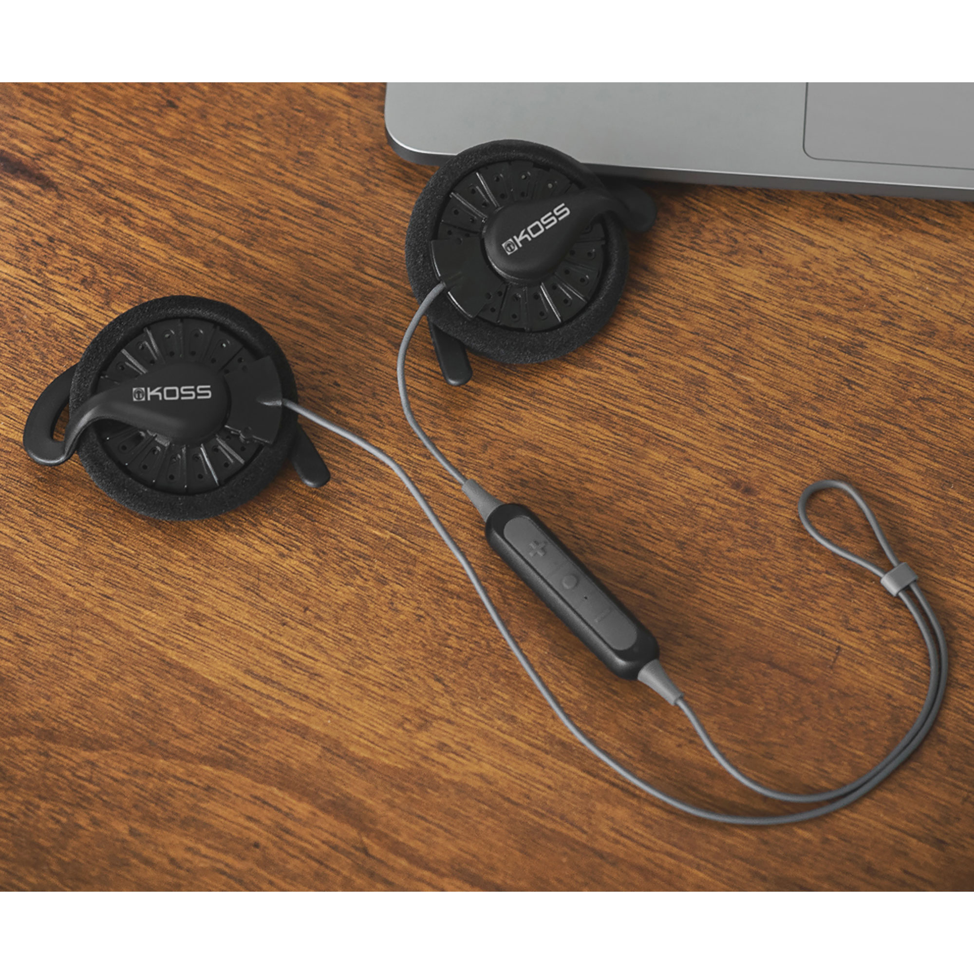 Czarne słuchawki KOSS z kablem i pilotem na drewnianym stole. W tle widoczny jest laptop.