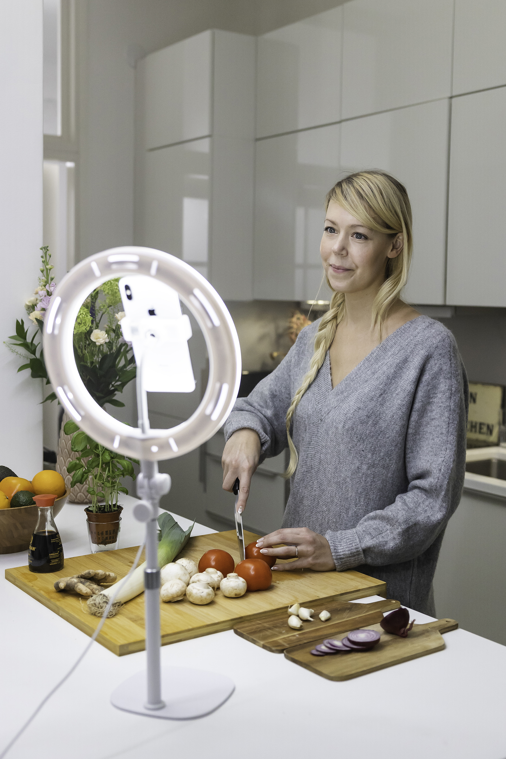 Eine Frau schneidet Tomaten mit einem Messer. Vor ihr befindet sich ein Ringlicht. Küchenumgebung mit Zutaten.