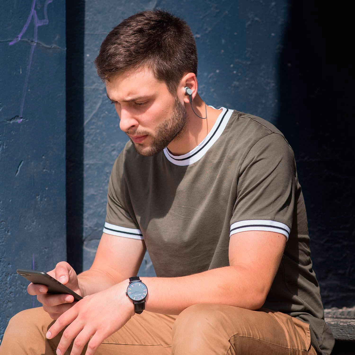 Un uomo seduto, guarda il suo telefono, indossa un orologio e auricolari. Lo sfondo è un muro blu.