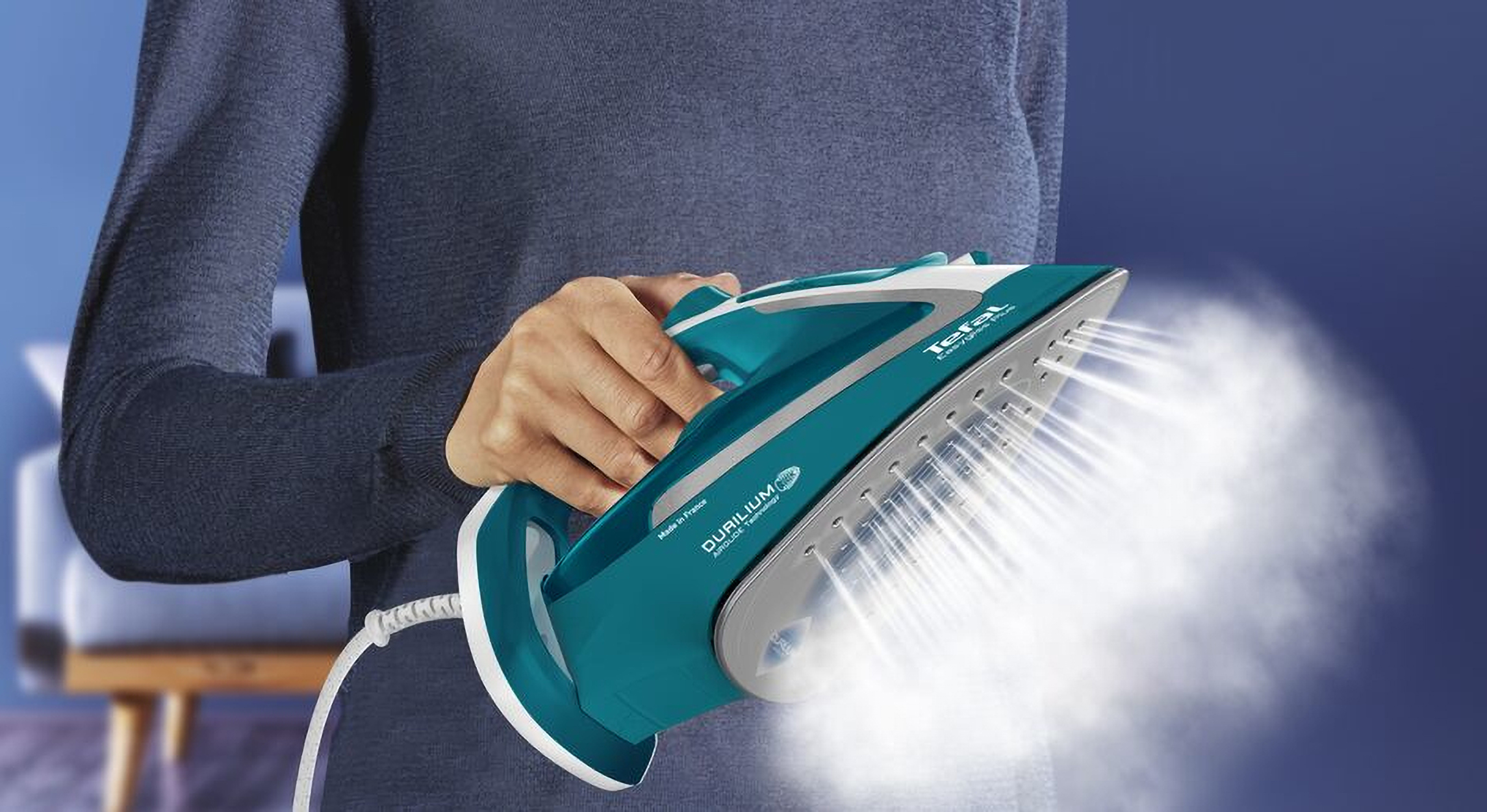 Una persona sostiene una plancha turquesa que emite vapor. Fondo azul, mango blanco.