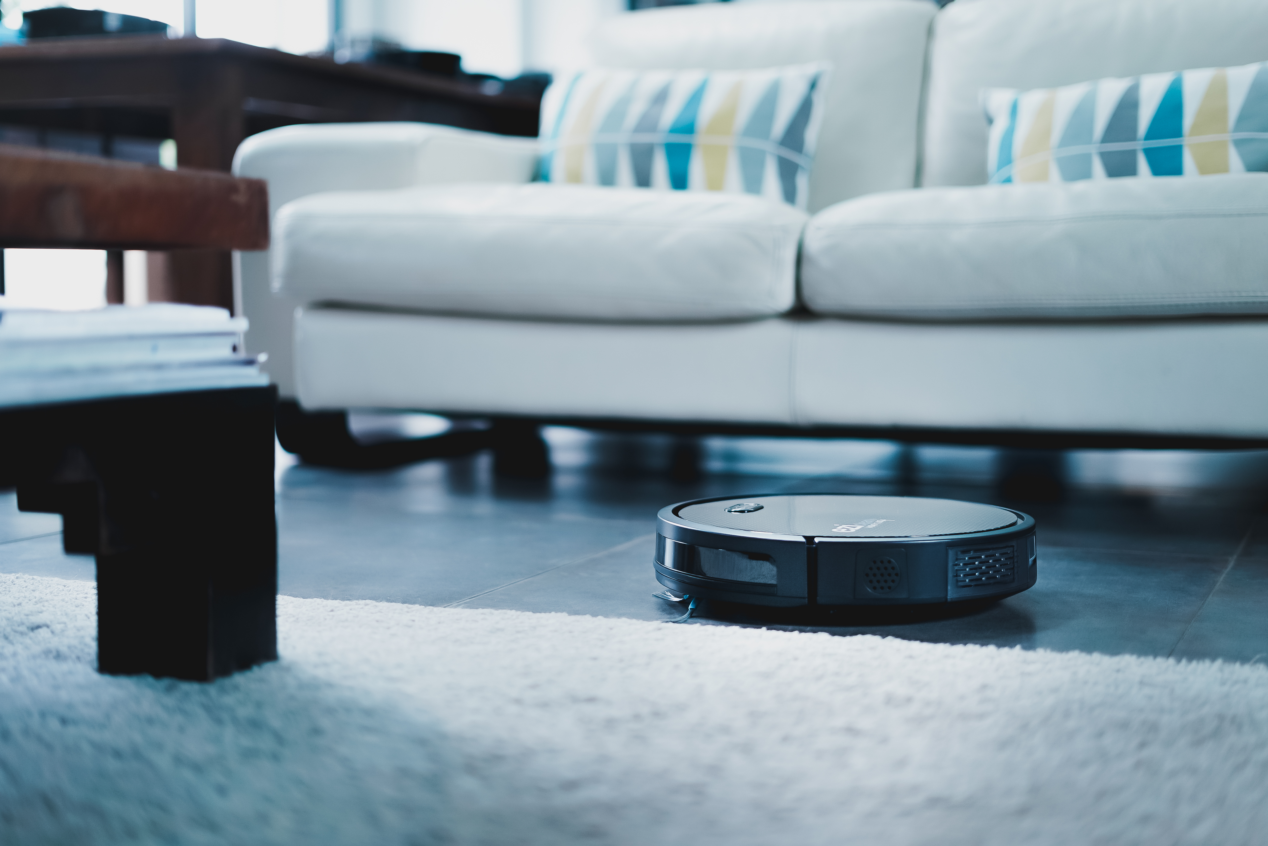 Una aspiradora robot negra está en el suelo en una habitación con un sofá blanco y una alfombra.
