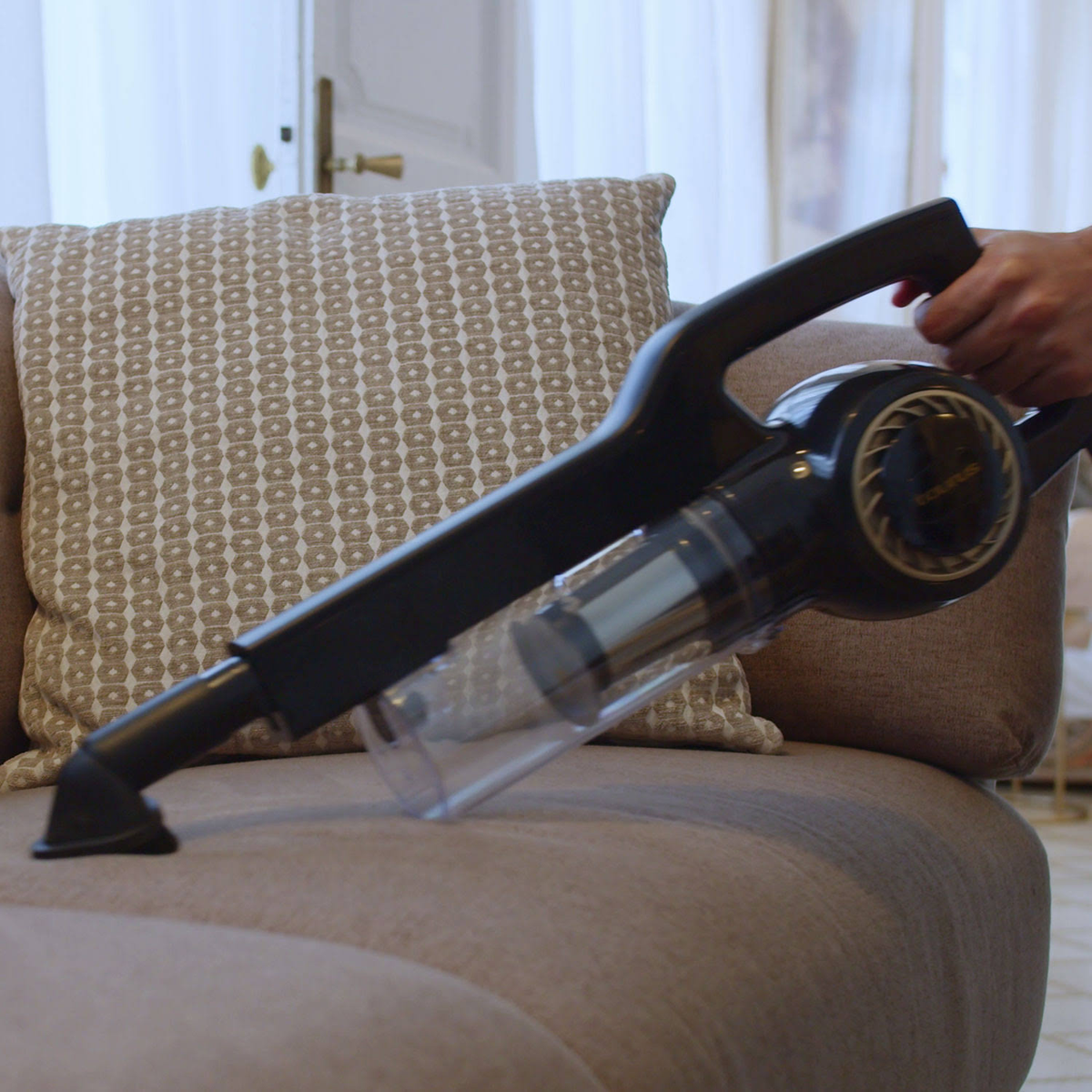 Osoba odkurza beżową sofę czarnym i przezroczystym odkurzaczem. Tło jest rozmyte.