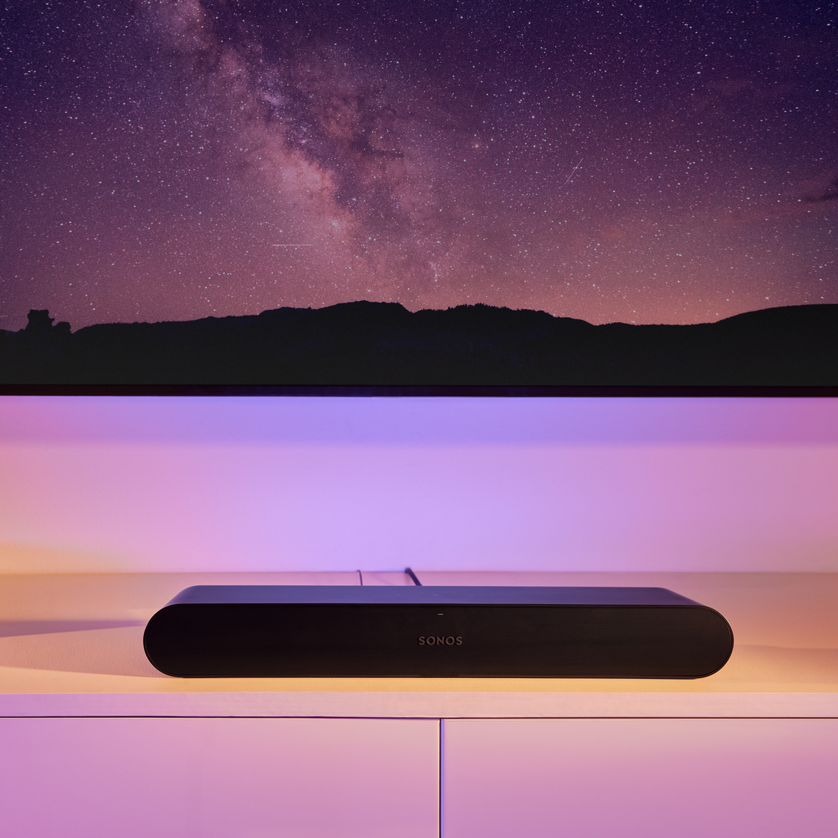 Schwarze Soundbar auf einem weißen Schrank unter einem Fernseher vor sternenklarem Nachthimmel.