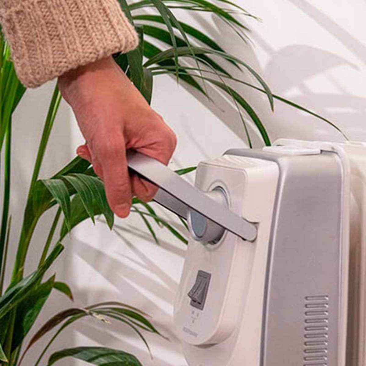 Dłoń pociąga za srebrną rączkę na białym grzejniku. Z tyłu zielone rośliny.