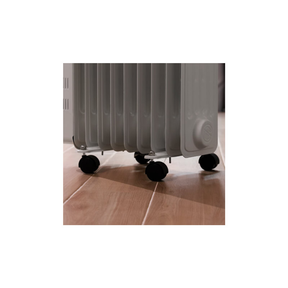 Szary grzejnik na kółkach. Drewniana podłoga. Biała ściana w tle. Skupienie na grzejniku.