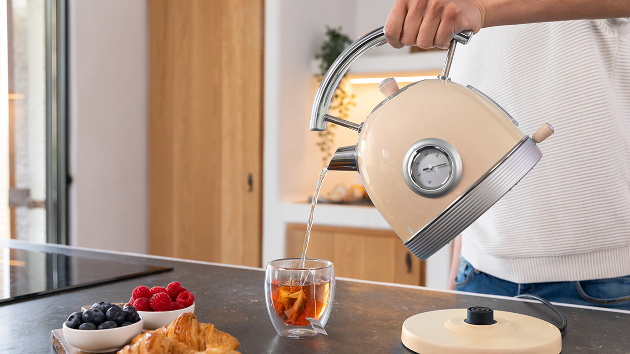 Osoba nalewa gorącą wodę z kremowego czajnika do szklanki herbaty na blacie kuchennym.