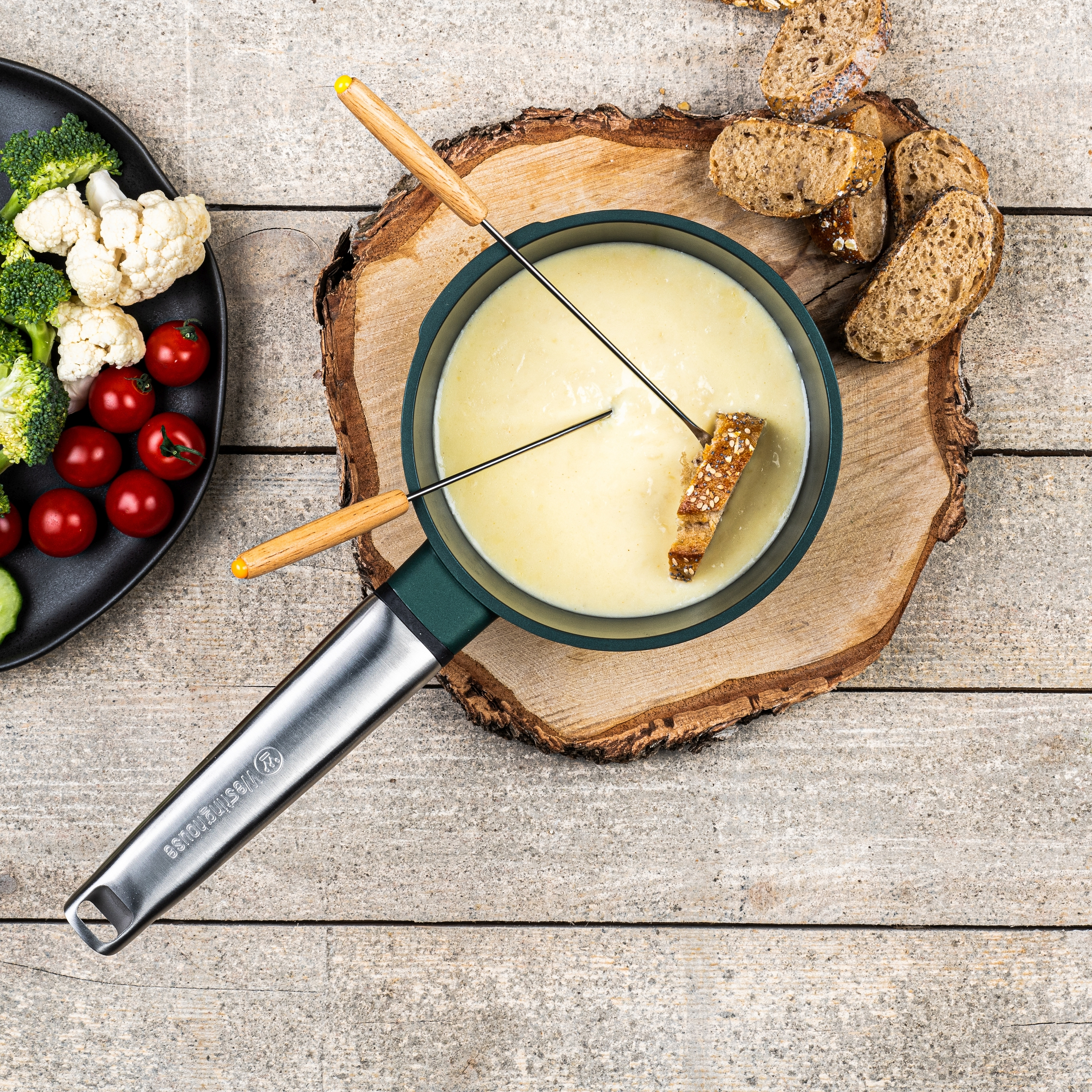 Fonduetopf auf Holz. Umgeben von Brot, Gemüse und Utensilien.