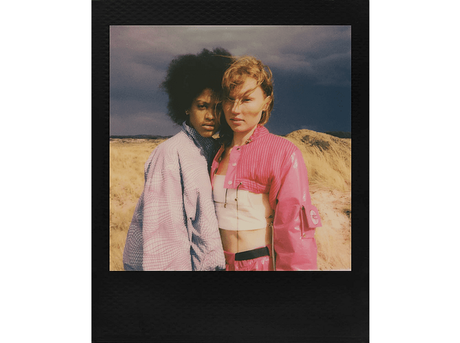 Foto Polaroid, dos mujeres. Una en rosa, una en gris. De pie en un campo.