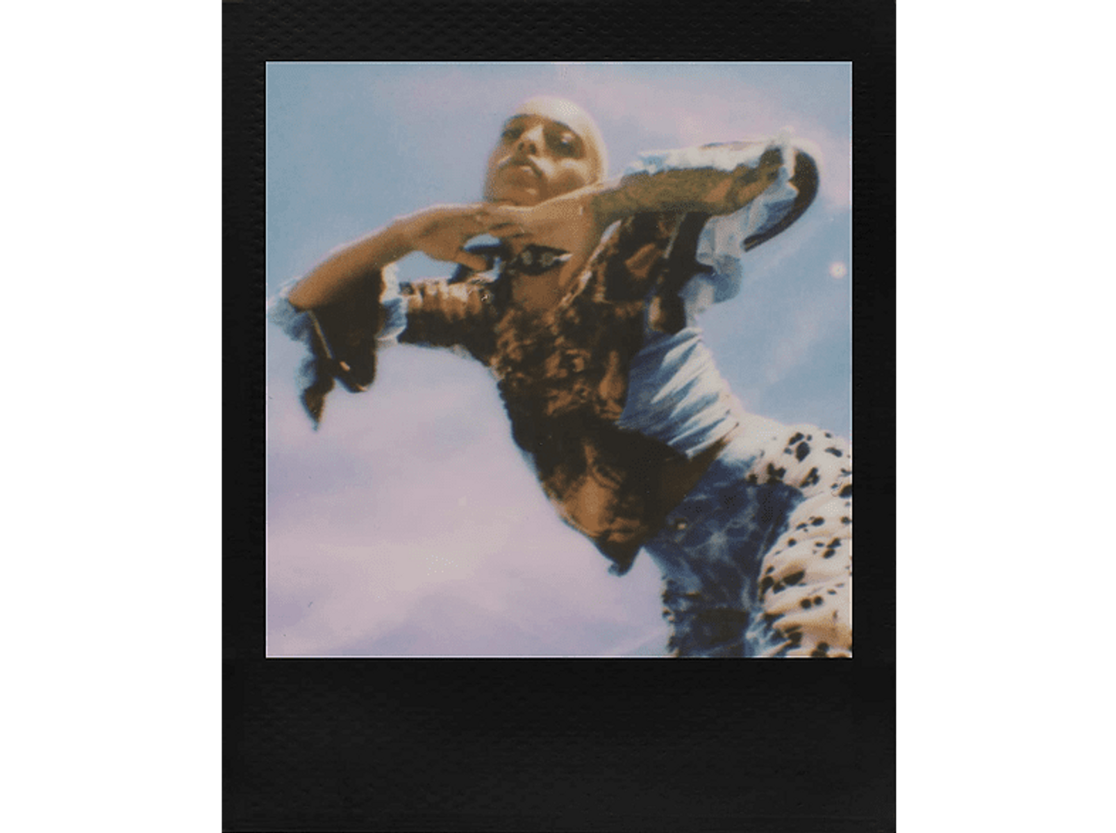 Foto Polaroid de una persona con ropa estampada, brazos levantados, contra un cielo nublado.