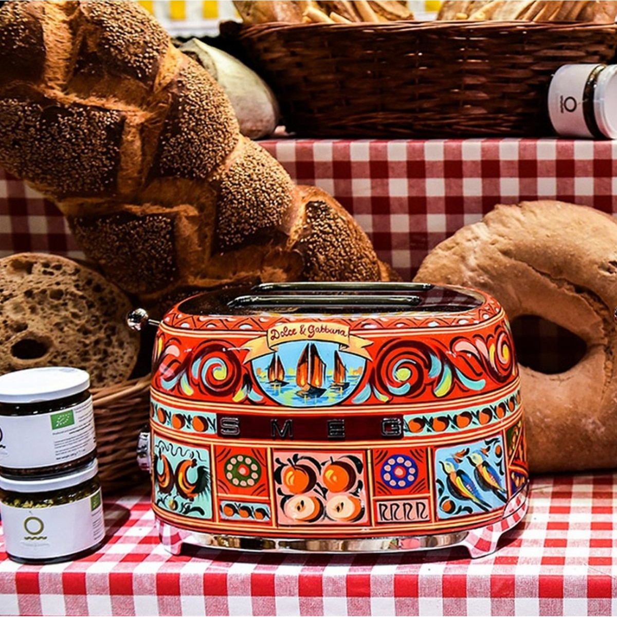 Una tostadora Dolce & Gabbana colorida sobre un mantel a cuadros rojo, con pan y frascos alrededor.