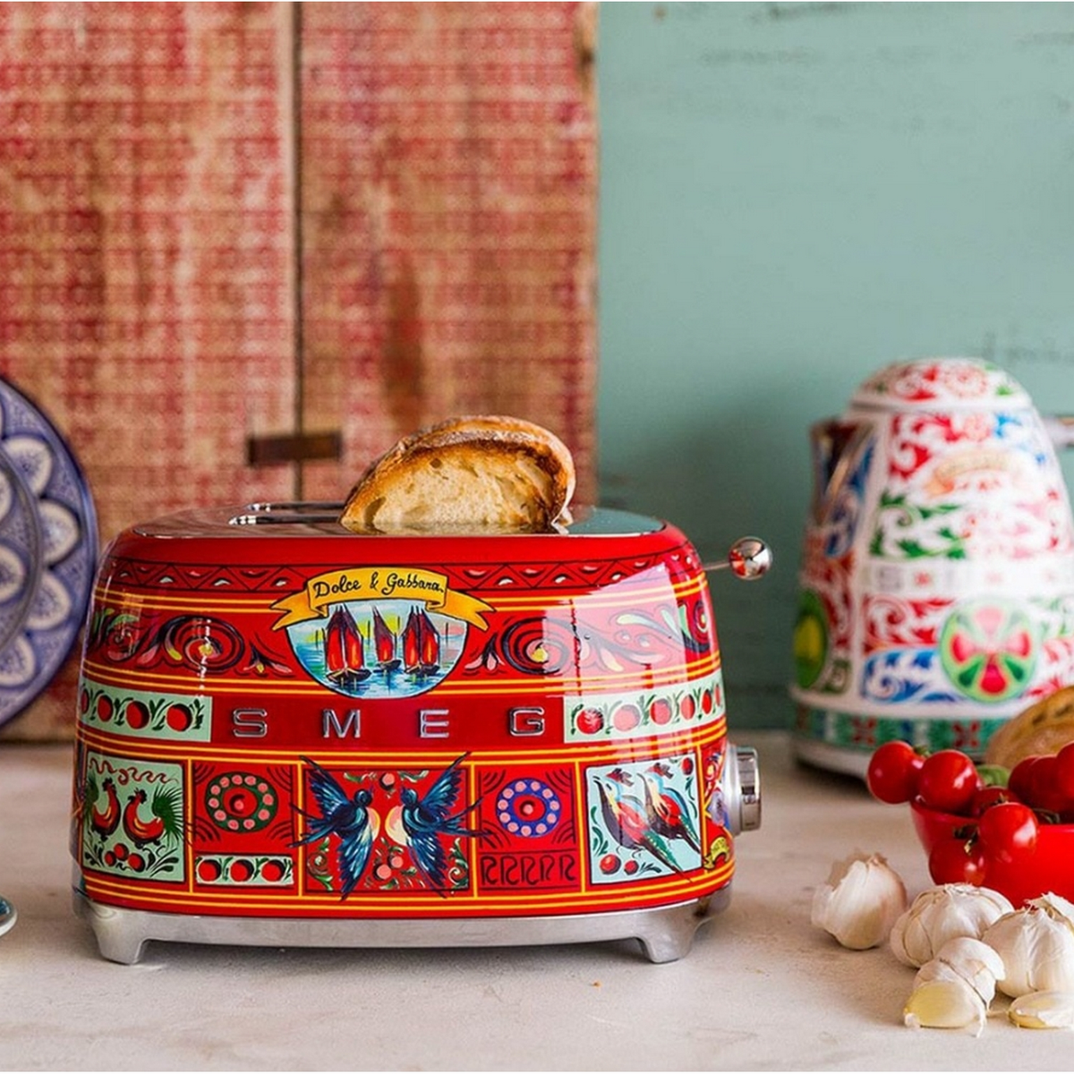 Tostadora SMEG roja con diseños coloridos. Una rebanada de pan sobresale. Hay otros elementos alrededor.