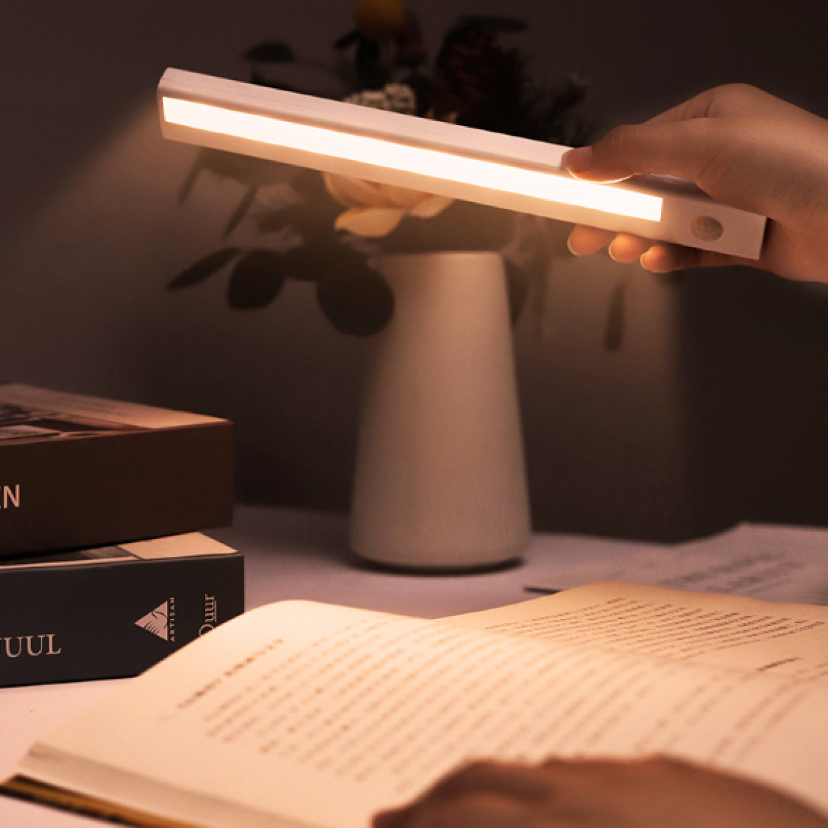 Een hand die een witte LED-lichtbalk boven een bureau houdt. Er liggen boeken, een vaas en een wazig open boek.