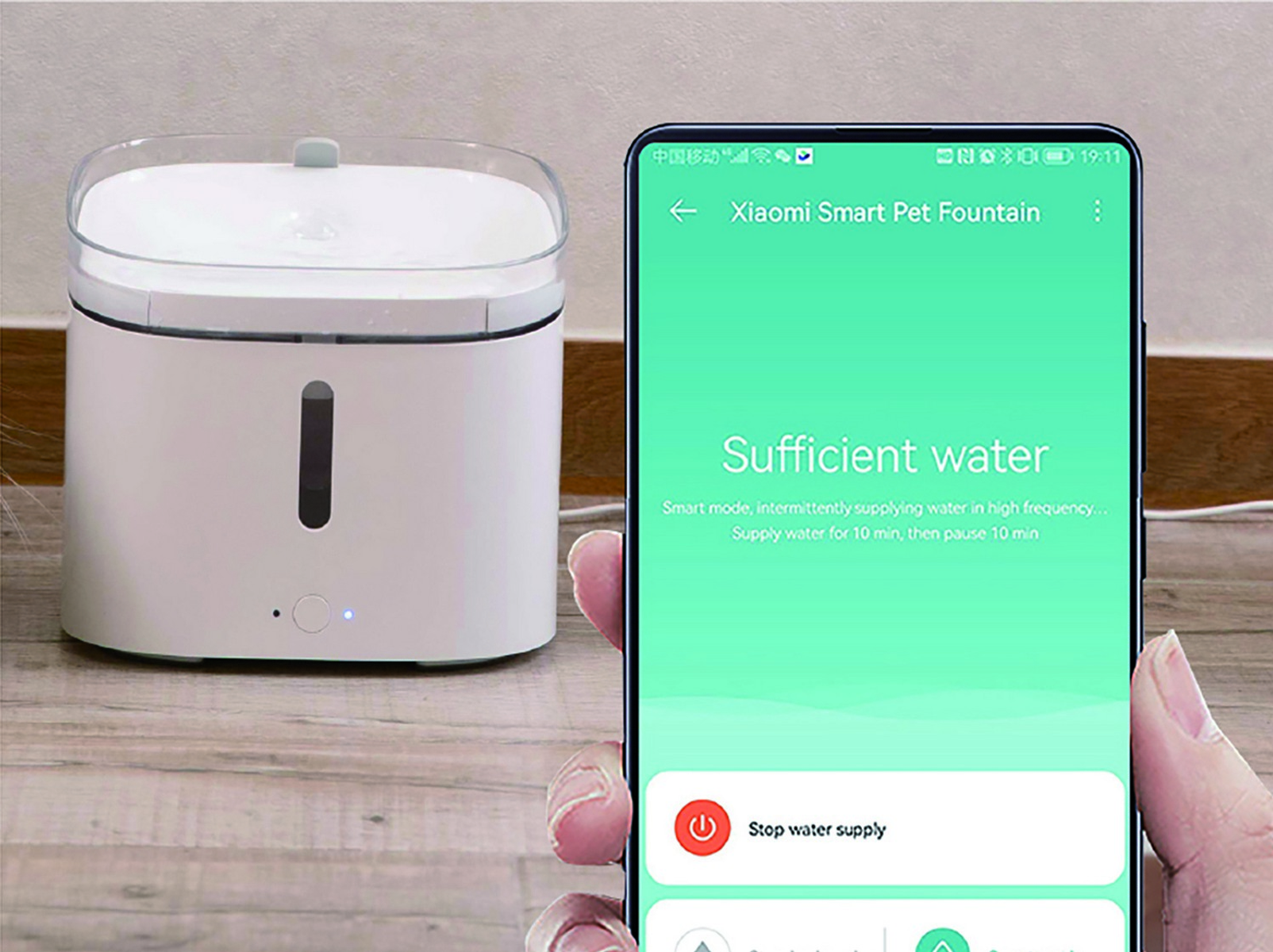 Ein Smartphone zeigt die Xiaomi-Haustierbrunnen-App. Der weiße Brunnen steht auf einer Holzoberfläche.