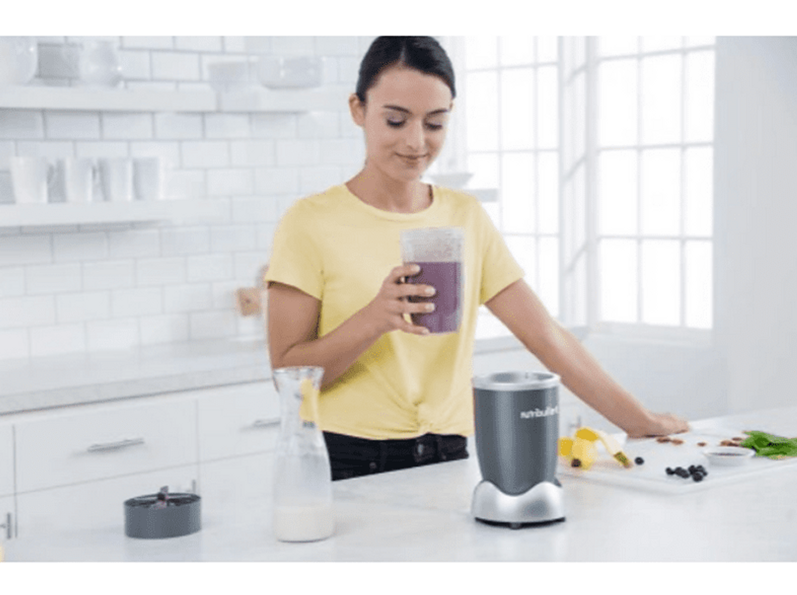 Mujer preparando un batido en la cocina. Licuadora, ingredientes y un vaso presentes.